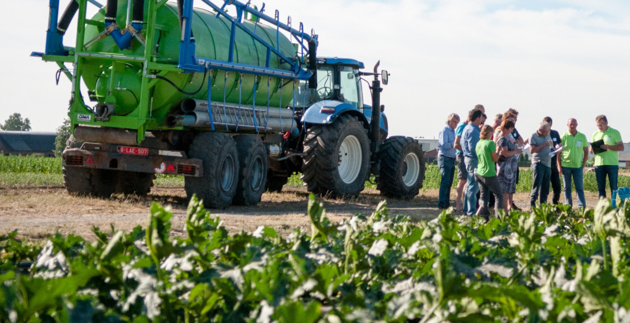 Photo of an agricultural vehicle in a field of crops with people standing nearby wearing high visibility clothing.