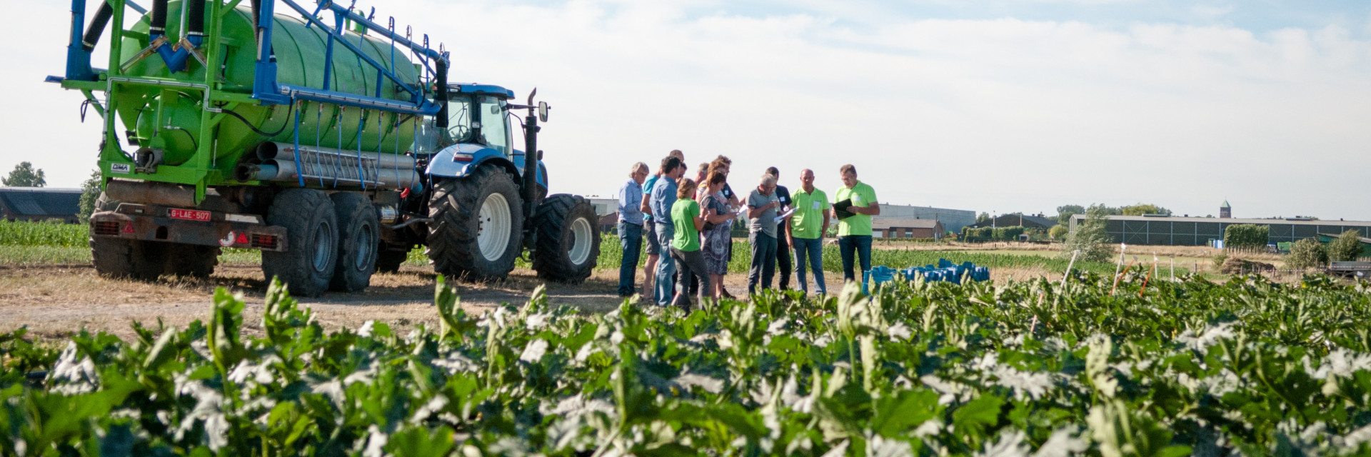 Photo of a field of crops with a large agricultural vehicle in the middle and a group of people wearing high vis jackets standing nearby.
