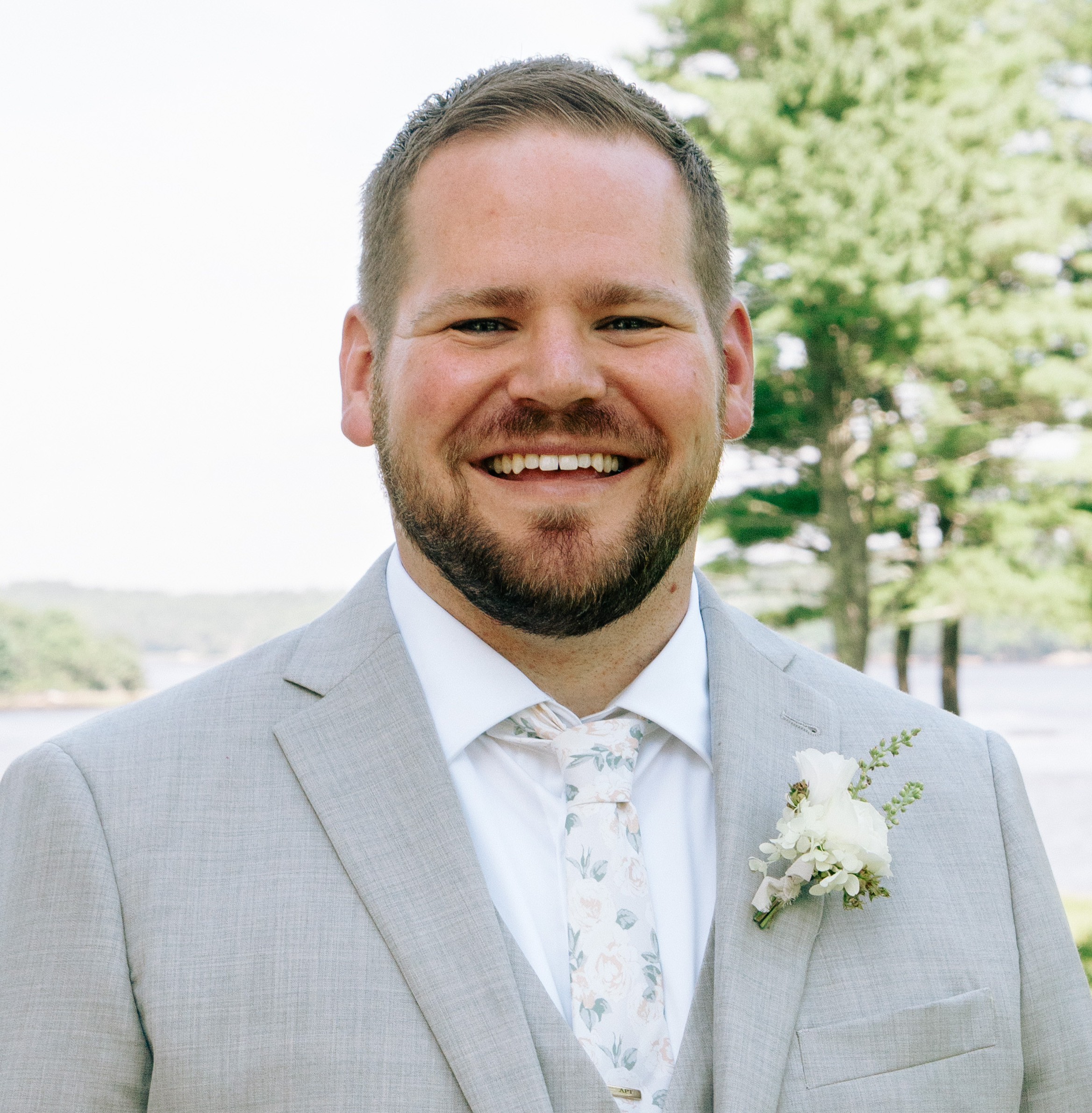 Photo of Andy Towne. He's wearing a pale coloured suit with a white shirt and patterned white tie, and a white flower arrangement in his button hole. He's a white man with short blonde hair and a beard and moustache. He's smiling at the camera with trees in the background.