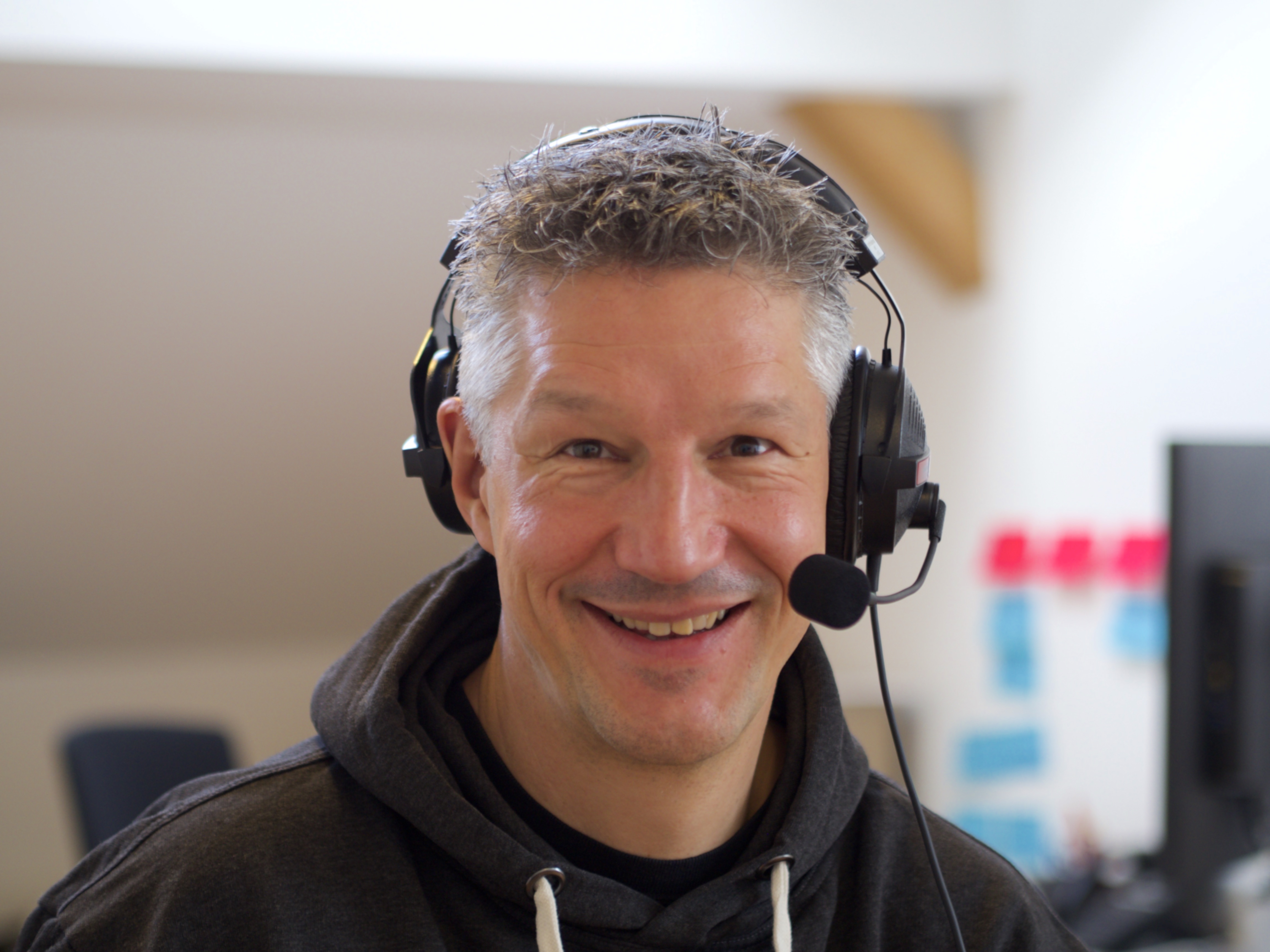 Photo of Ekke. He has short grey hair and is wearing a headset with a microphone and is smiling at the camera. He is wearing a black hoodie with white strings and is photographed in an office environment.