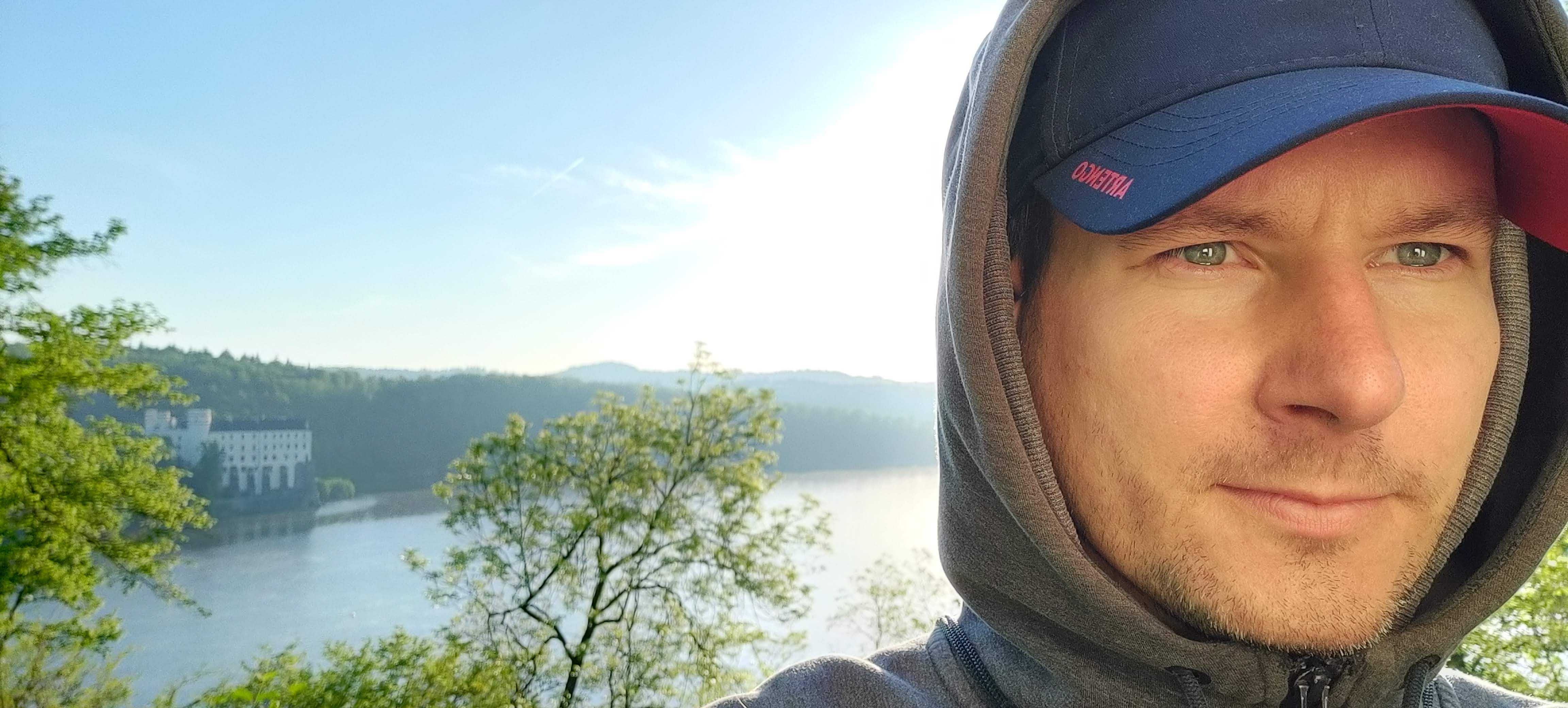 Photo of John Linhart in front of a large expanse of water with trees on either bank. He's a white man and is clean shaven, wearing a peaked cap and a hoodie with the hood up and is looking past the camera to the right.