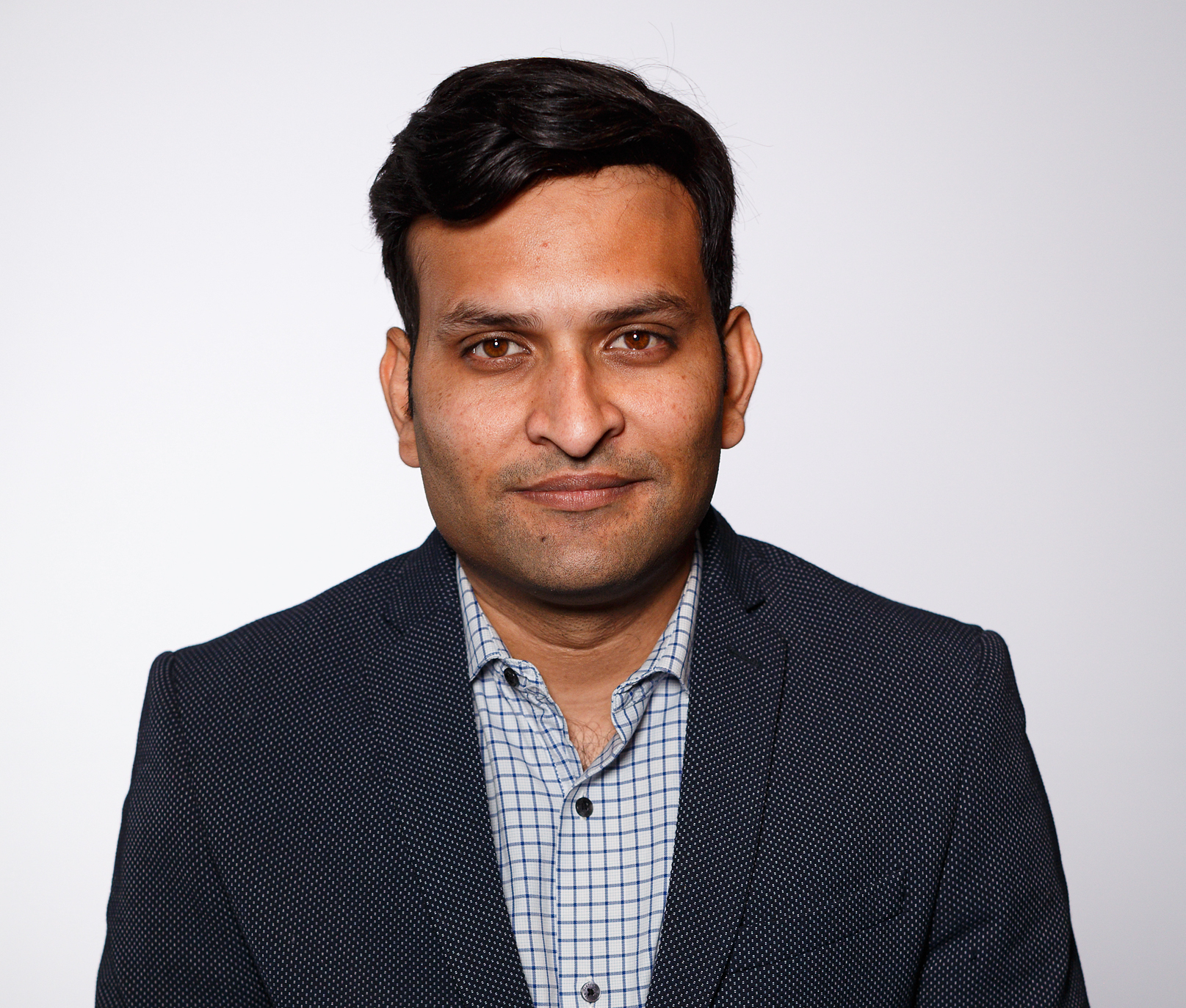 A photo of Prateek, he's an Indian man, clean shaven with dark mid-length hair wearing a black blazer and a checked white and blue shirt. He is smiling at the camera and is photographed against a white background.