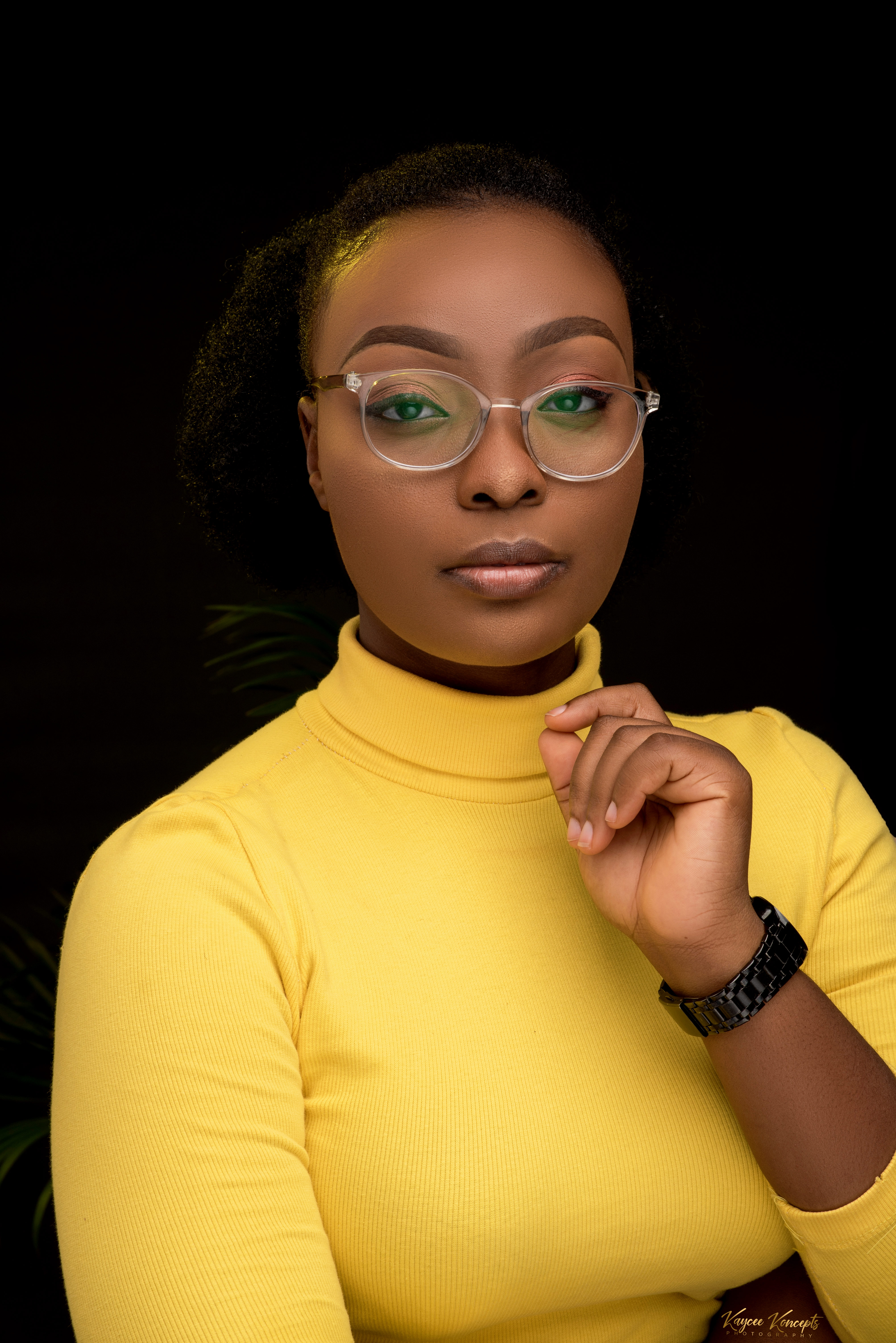 A photo of Favour, she is a black woman wearing a yellow polar neck jumper and clear-framed glasses with her hair tied back. She has her left hand raised to her face and is photographed against a black background.