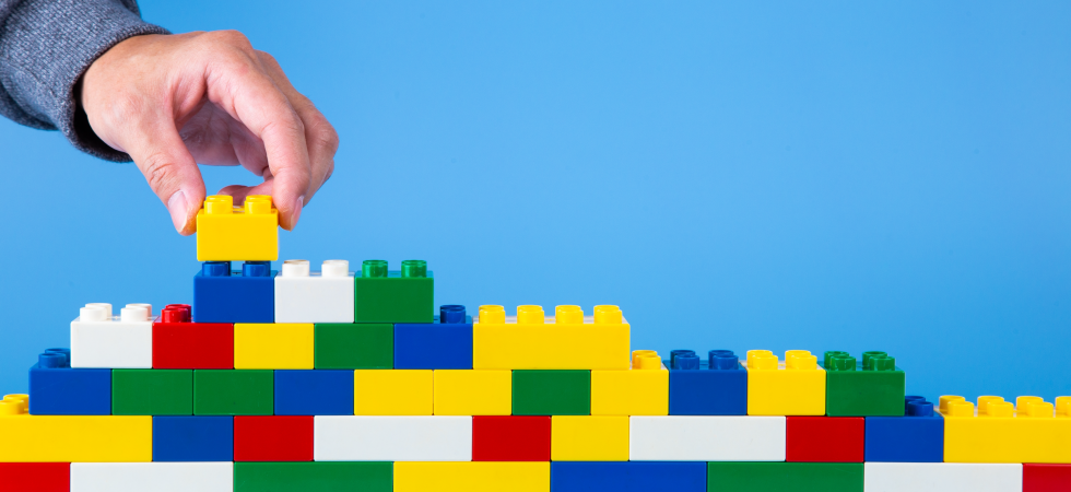 Photo of lego bricks of different colours stacked up with a hand adding another brick, against a blue background.