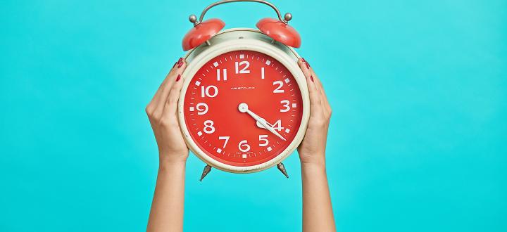 Photo of a red alarm clock being held by two hands on a cyan background