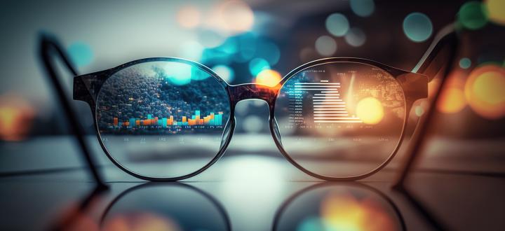 Photo of a pair of glasses on a table - in the glasses you can see charts and graphs and the background has blurred lights.