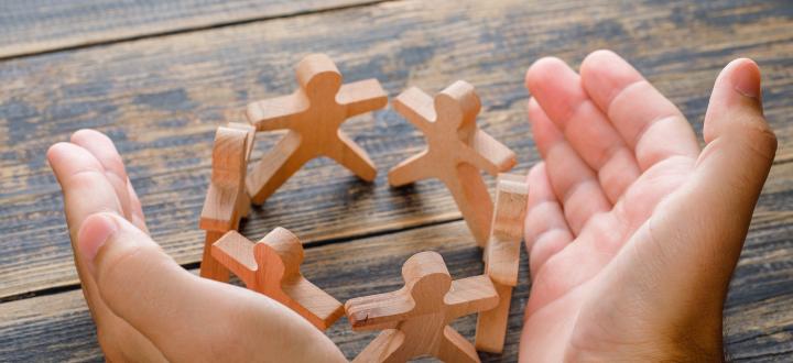 Photo of hands surrounding wooden cutout figures who are all in a circle, on a wooden bench.