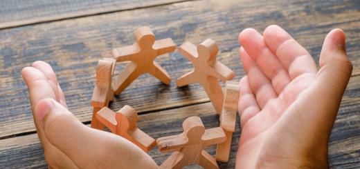 Photo of hands surrounding wooden cutout figures who are all in a circle, on a wooden bench.