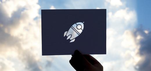 A blue cloudy sky with sun shining through the clouds, and a dark hand holding a black piece of card with a rocket ship cut out in the middle.