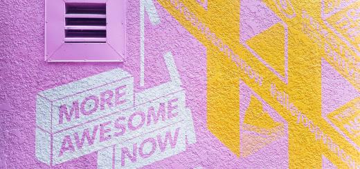 A wall painted bright pink and yellow with 'more awesome now' stencilled on a white background in pink paint